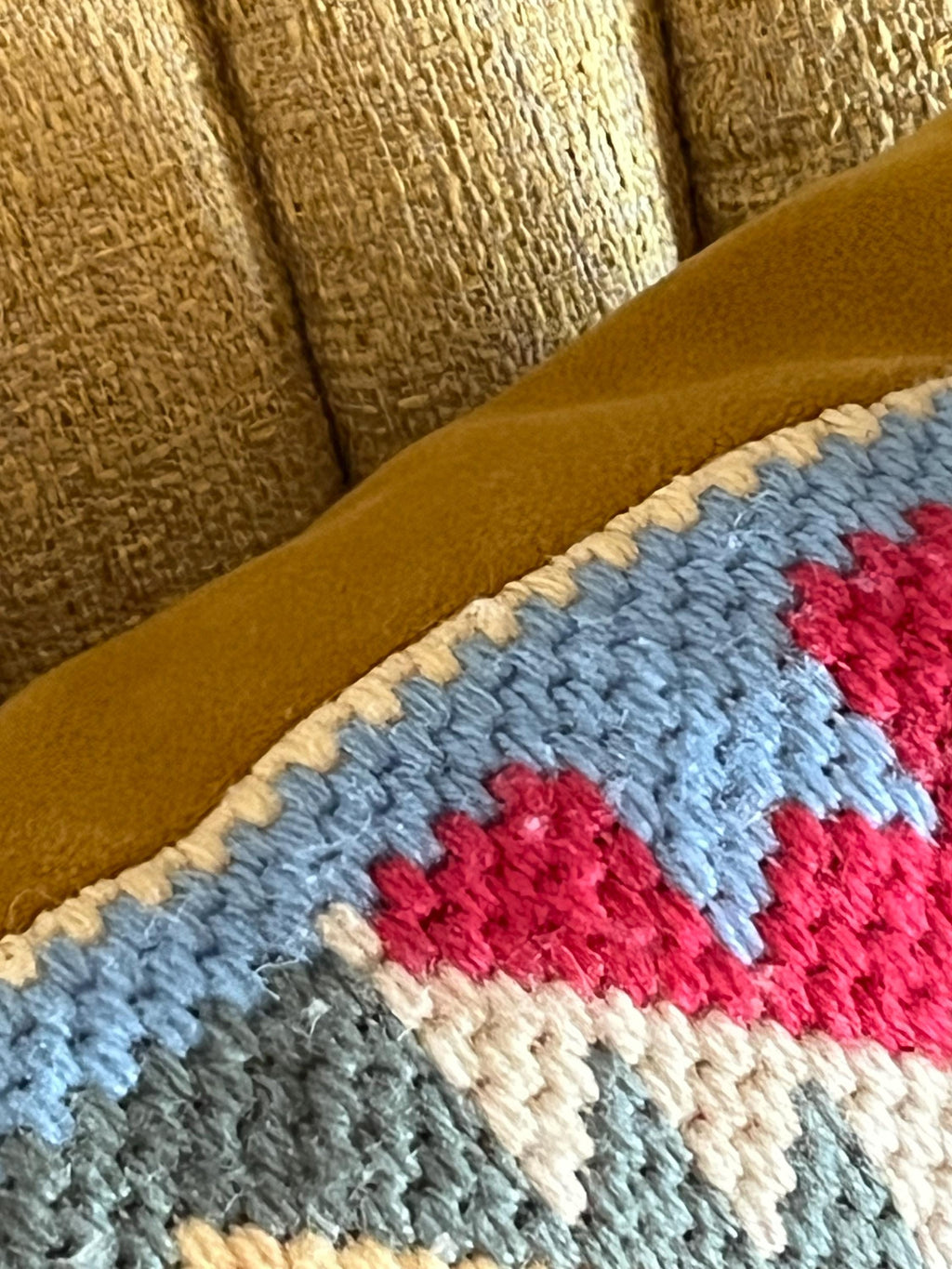 A close-up of a wool tapestry with a geometric pattern in light blue, bright pink, cream, and gray, resting against a mustard yellow fabric surface on a textured beige upholstered chair.