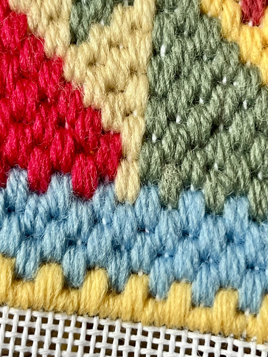 Close-up of a colorful wool tapestry with textured rows in red, cream, sage green, light blue, and yellow, with a white mesh backing visible along the bottom edge.